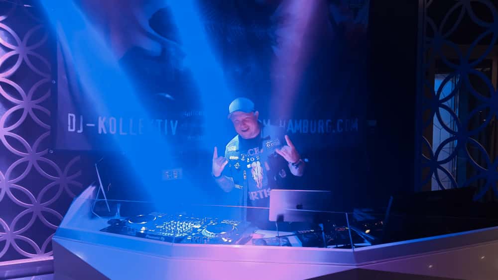 DJ Borncore hinter den Decks mit DJ-Kollektiv-Banner, Hörner oben.